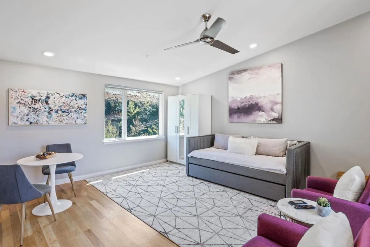 A cozy, modern space featuring a daybed near a large window, abstract artwork, and a small dining table. The ample natural light and comfortable seating create a welcoming environment for unwinding after a day of city adventures or sharing a quiet moment with a loved one while watching the sunset.