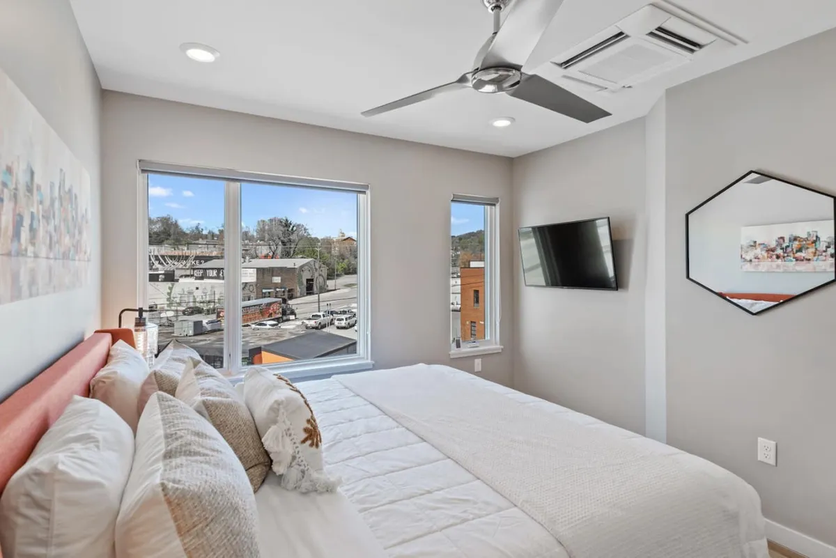 A comfortable bedroom with a cozy bed adorned with decorative pillows, a wall-mounted TV for evening relaxation, large windows offering a pleasant view, and stylish decor. The mood is modern yet inviting—ideal for unwinding after a busy day.