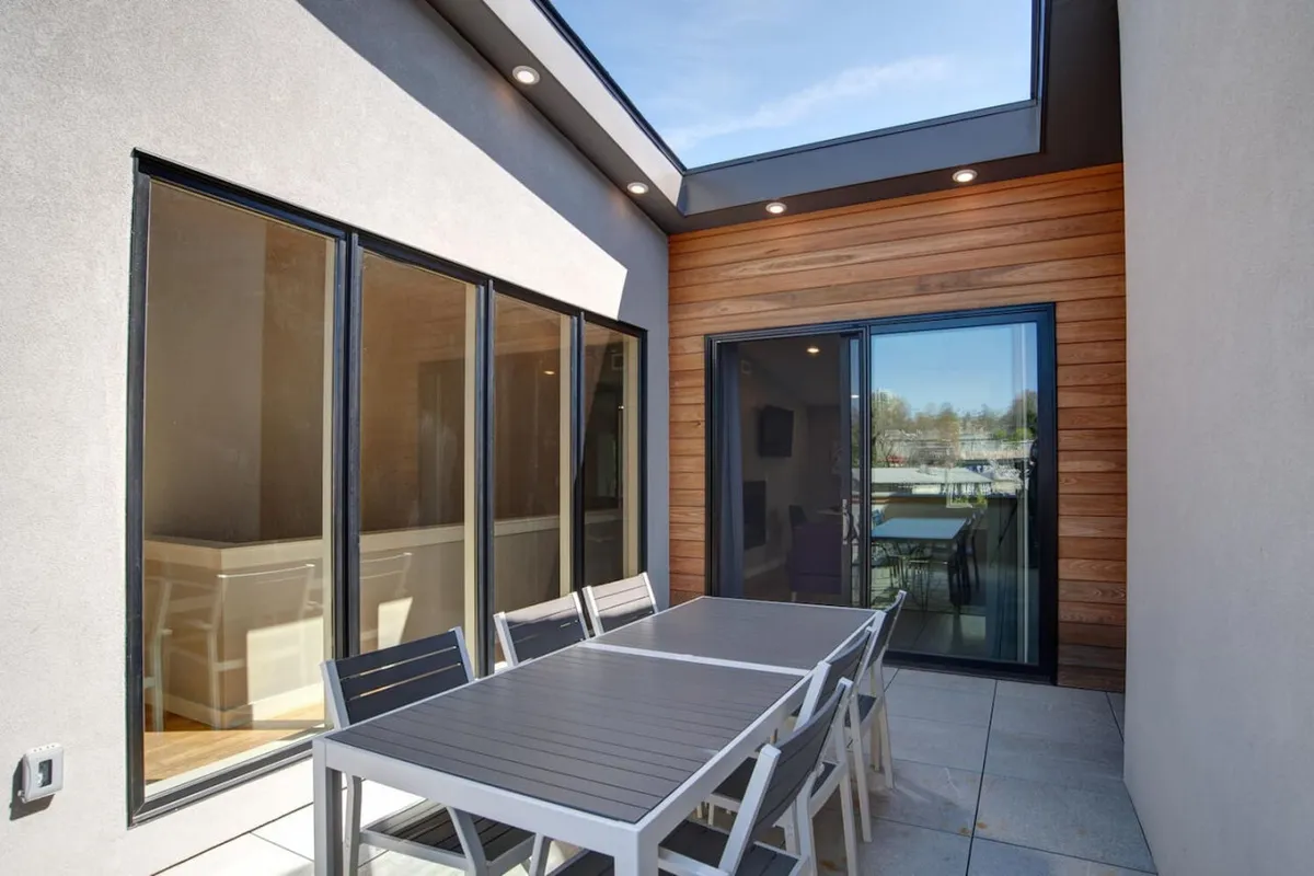 This image captures an inviting exterior dining area featuring a modern outdoor table set against large windows and sleek wooden paneling. Sunlight floods the space, creating a welcoming atmosphere ideal for both daytime relaxation and evening gatherings. The design suggests a seamless connection between the indoor living areas and the surrounding urban environment, embodying the lifestyle of stepping out into vibrant city life.