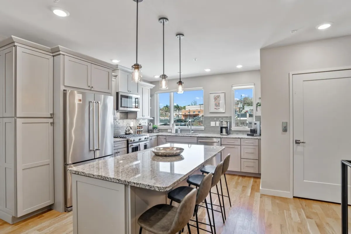 A contemporary kitchen features a large granite-topped central island, stainless steel appliances, and expansive windows letting in natural light. The space is designed for both culinary creativity and socializing, with room for guests to gather around the island or enjoy meals together.
