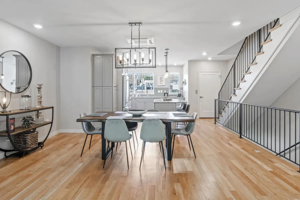 A spacious dining area with a large table set beneath a modern chandelier, adjacent to an elegant staircase and a decorative mirror. The setting invites group meals or celebratory toasts before or after an evening out.