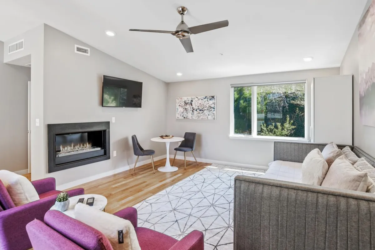 A modern, cozy interior scene with a plush sofa, a stylish gas fireplace, a table with chairs, and vibrant artwork on the wall. Natural sunlight brightens the room, creating an inviting lounge atmosphere perfect for relaxing, entertaining, or unwinding by the fire.