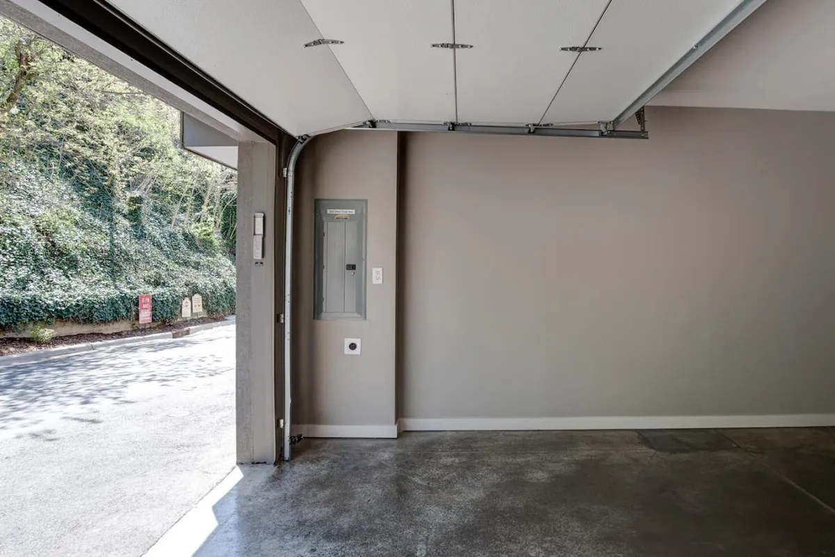 A clean, functional garage with an open garage door, concrete floor, and wall-mounted electrical panel. Ample space for double parking and visible entry point into the home evoke convenience and security.