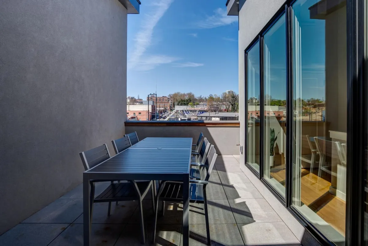 This image captures an inviting rooftop terrace with an outdoor dining table set against expansive city views. Large glass windows connect the exterior space with the interior, and the scene is set for relaxed socializing or enjoying a meal al fresco. The atmosphere is relaxed and social, ideal for evening gatherings or sunset cocktails.