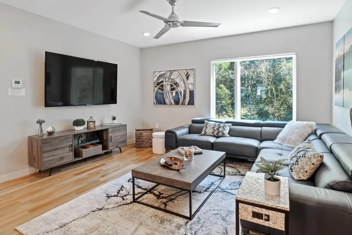 A stylish living room boasts a large sectional sofa facing a coffee table and a wall-mounted TV, with tasteful artwork and expansive windows streaming in daylight. The setting is ideal for relaxing after a day exploring the city or hosting movie nights with friends.