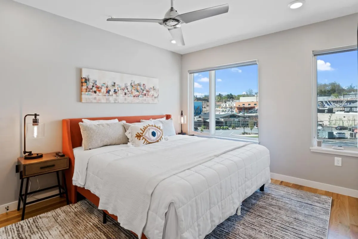 A serene bedroom featuring a plush bed with an orange headboard, modern artwork above the bed, and expansive windows that bathe the space in natural light. The room feels fresh, calm, and thoughtfully designed, inviting relaxation and comfort.