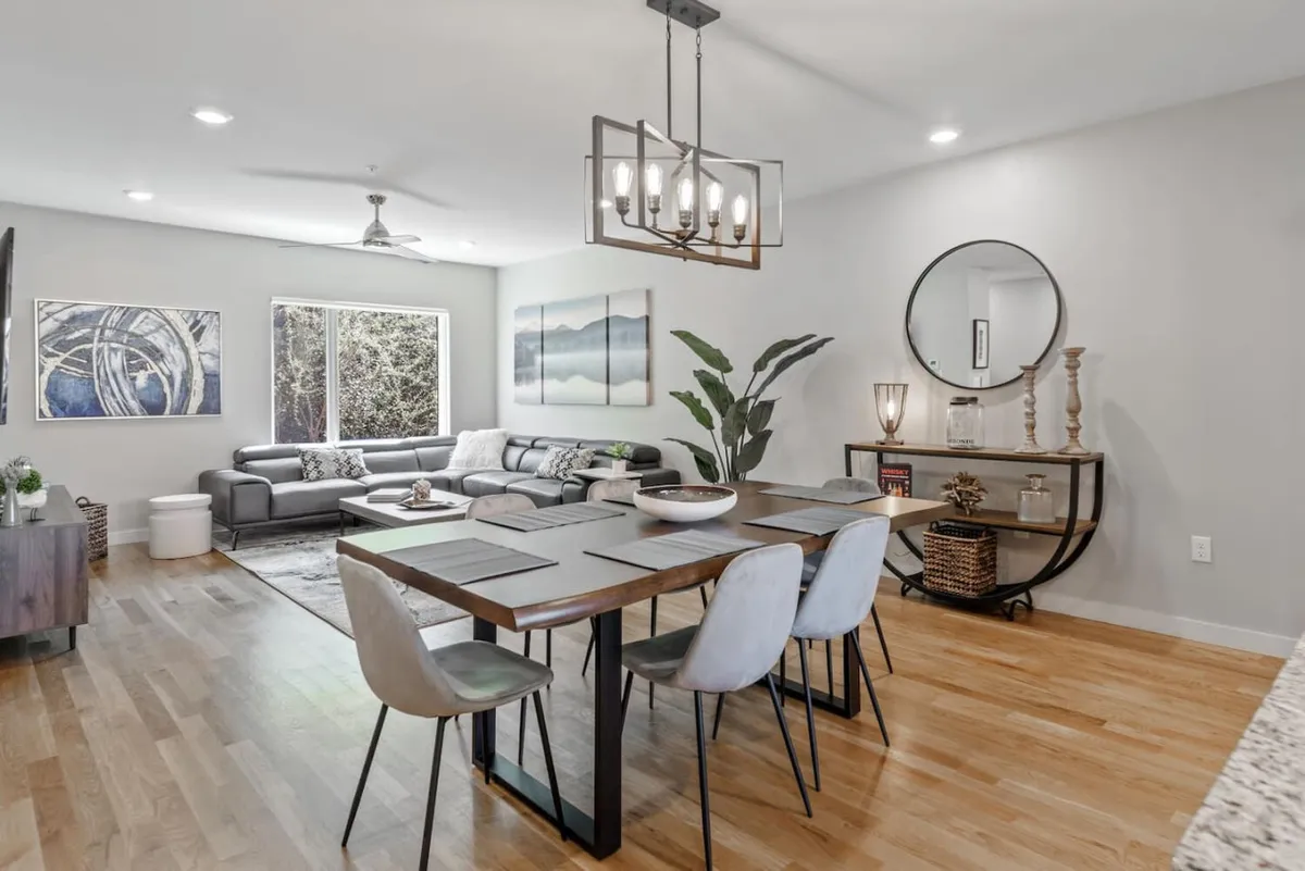 A contemporary, cozy open-plan living and dining space with a modern dining table set for two, a comfortable sofa, stylish artwork adorning the walls, and a large window that hints at city views outside. The overall atmosphere suggests both intimacy and urban sophistication, perfect for romantic dinners or relaxed evenings.
