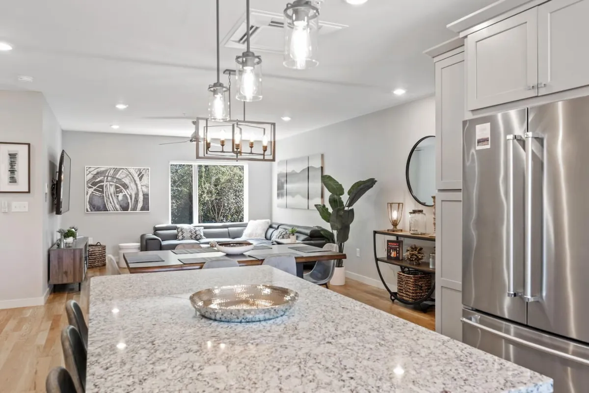 A contemporary open-plan kitchen and living area with a large granite island, stylish light fixtures, comfortable seating, and a touch of greenery. The space is warm and inviting, ideal for groups to unwind, share drinks, or enjoy late-night conversations after a night on the town.