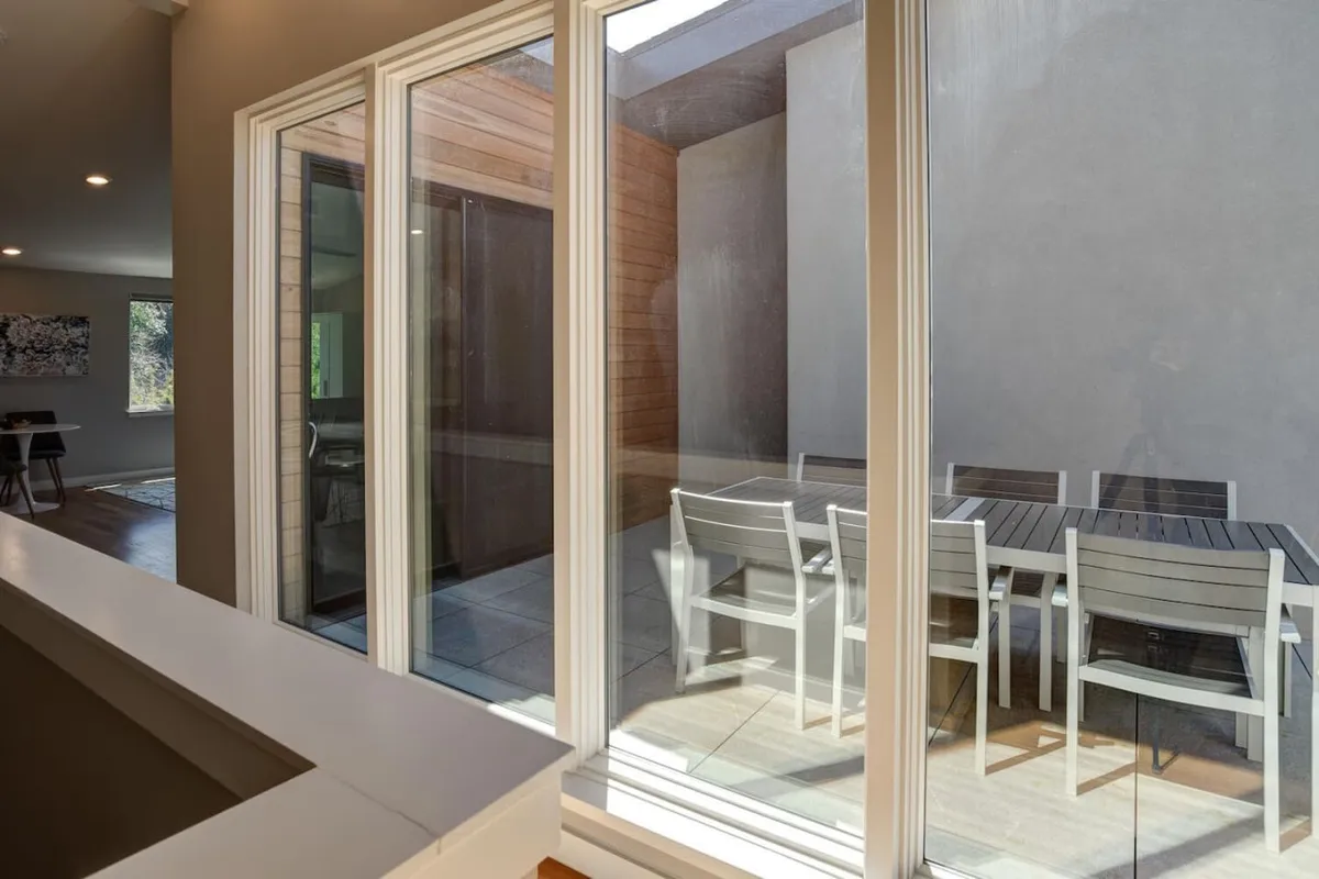 A view from inside looking out onto a stylish outdoor dining set, framed by expansive windows. Natural light fills both the interior and patio space, highlighting the fluid boundary between indoor comfort and the lively, walkable neighborhood beyond.